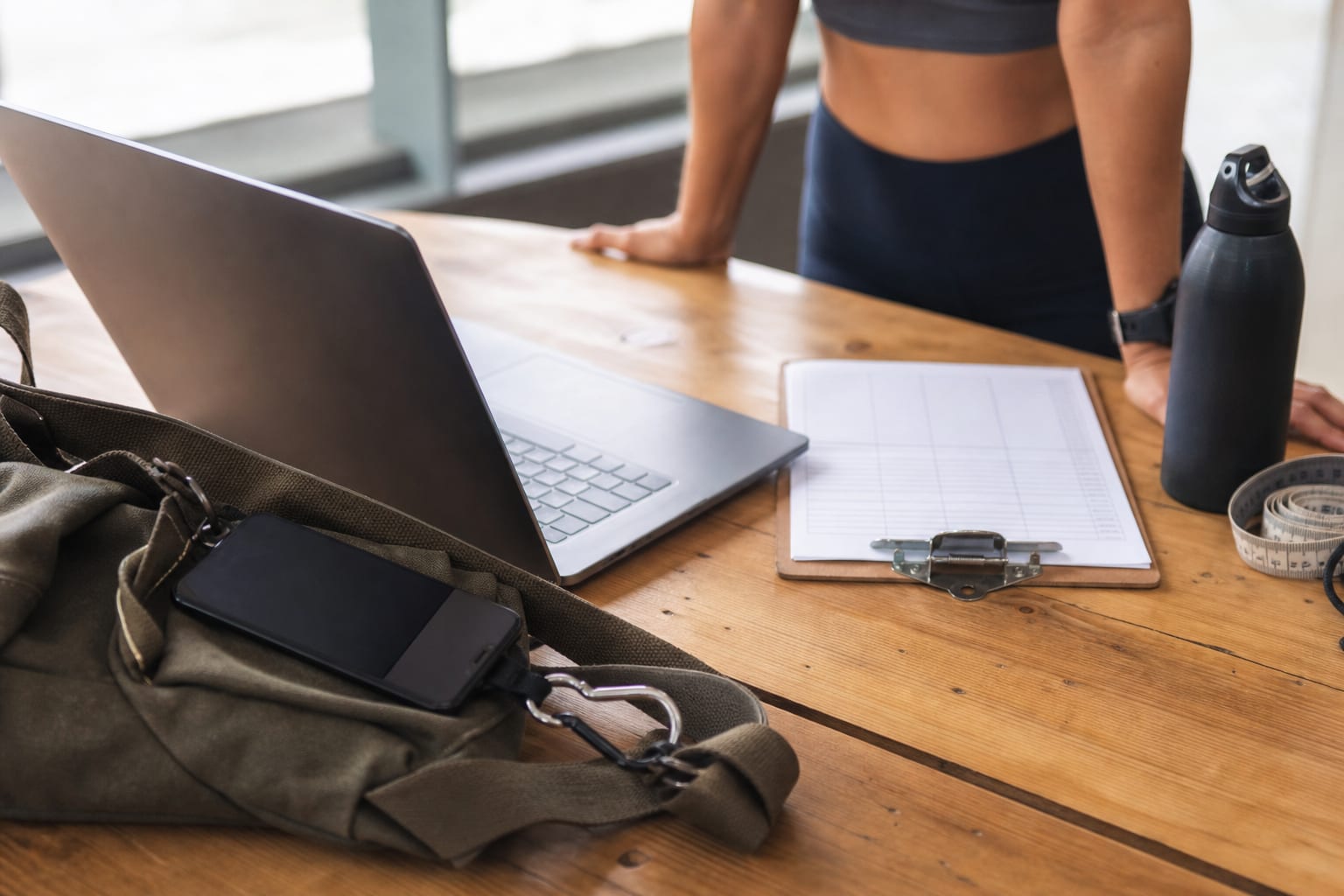 Bancada de recepção de estúdio com notebook, ficha de treino impressa e celular ao lado das mãos de uma professora