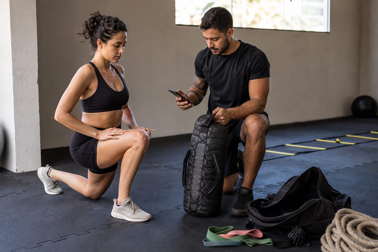 Personal trainer atendendo aluna no leg press de estúdio pequeno enquanto confere a série no celular